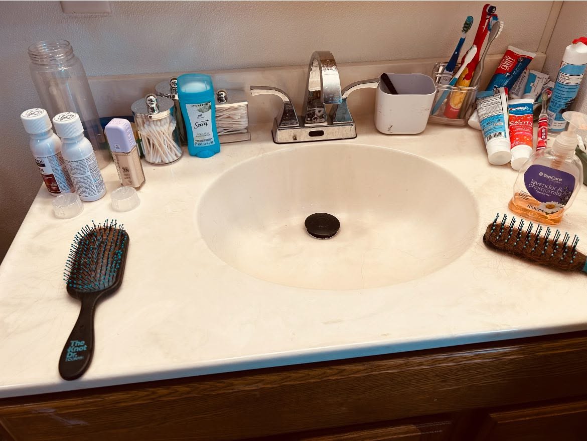 Bathroom vanity crowded with brushes and toiletries before cleaning